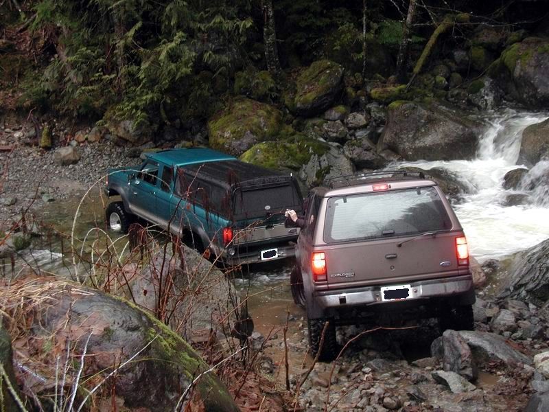 Teal pickup truck and brown SUV driving through a rocky stream.