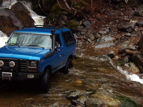 1985 Ford Bronco - A blue Ford Bronco is driving through a shallow, rocky stream with a waterfall in the background. The vehicle has off-road tires, a snorkel…