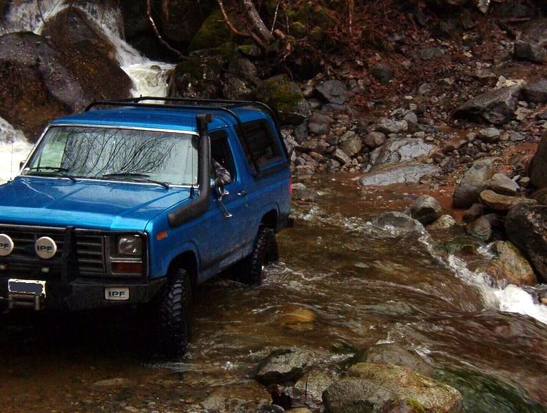 Blue Ford Bronco driving through a rocky stream with a waterfall.