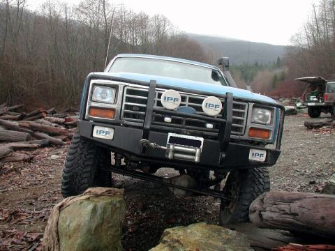 1985 Ford Bronco - A blue 1980s Ford Bronco is parked on a rocky, off-road trail with logs scattered around. The vehicle features a custom front bumper with I…