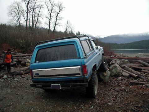 1985 Ford Bronco - The rear-left quarter of a blue Ford Bronco is shown parked on a rocky, debris-strewn area next to a body of water. The vehicle is angled,…