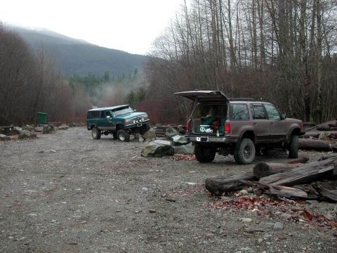1985 Ford Bronco - Two modified off-road SUVs are parked on a gravel lot next to a forest. The teal SUV on the left is angled with its front wheels on rocks,…