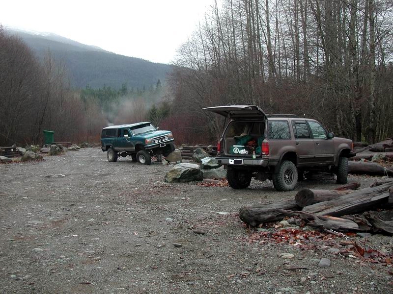 Two modified SUVs parked on a gravel lot near a forest, one with its hatch open.