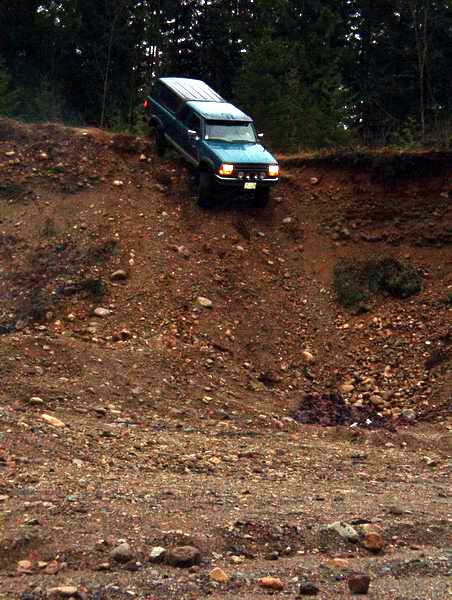 1985 Ford Bronco - A teal Ford Bronco with a white camper shell is driving down a steep, rocky dirt embankment with trees in the background.