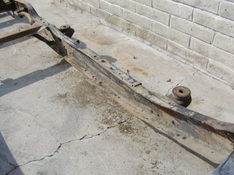 1981 Ford Bronco - Close-up of a bare, surface-rusted boxed frame rail on a bare chassis sitting on a cracked concrete driveway next to a white block wall. Tw…