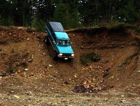 1985 Ford Bronco - A teal Ford Bronco with a black camper shell is navigating a steep, rocky incline in a wooded area. The vehicle is angled downwards on the…