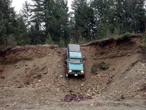 1985 Ford Bronco - A teal Ford Bronco is navigating a steep, rocky incline in a wooded area. The vehicle is angled downwards into a gully, with trees and erod…