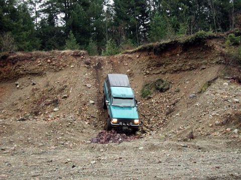 1985 Ford Bronco - A teal-colored SUV is navigating a steep, rocky incline in a wooded area. The vehicle is positioned at an angle, descending into a narrow g…