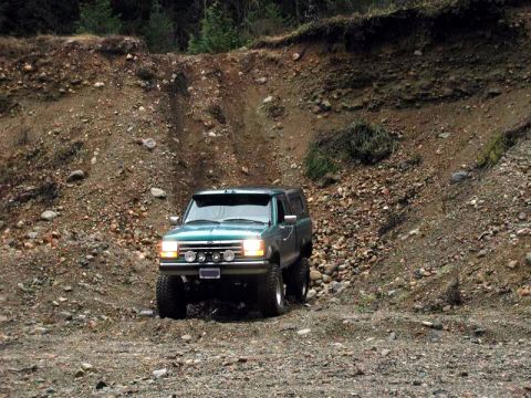 1985 Ford Bronco - A teal Ford Ranger pickup truck with a camper shell is driving on a rocky, uneven terrain with a steep dirt embankment in the background.
