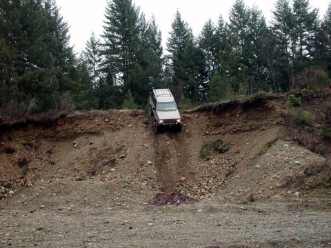 1985 Ford Bronco - A tan SUV navigates a steep, rocky incline with a wooded area in the background. The vehicle is positioned on the slope, descending into a…