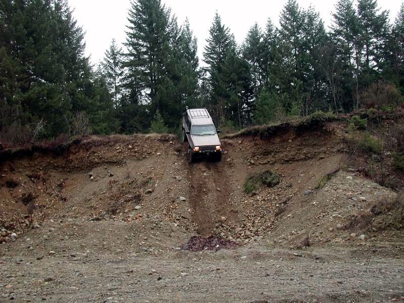 Tan SUV driving down a steep, eroded dirt and rock slope with trees in the background.