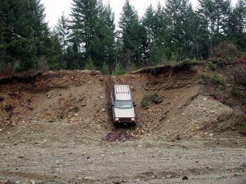 1985 Ford Bronco - A tan SUV is driving down a steep, rocky embankment in a wooded area.