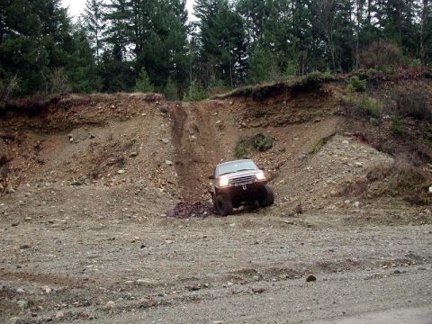1985 Ford Bronco - A lifted, dark-colored SUV is navigating a steep, rocky incline next to a dirt embankment. The vehicle appears to be off-roading in a woode…