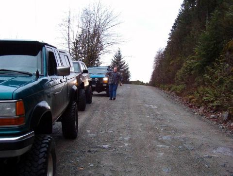1985 Ford Bronco - A line of lifted off-road vehicles, including a teal Ford Ranger in the foreground, are parked on a gravel road next to a wooded hillside.…