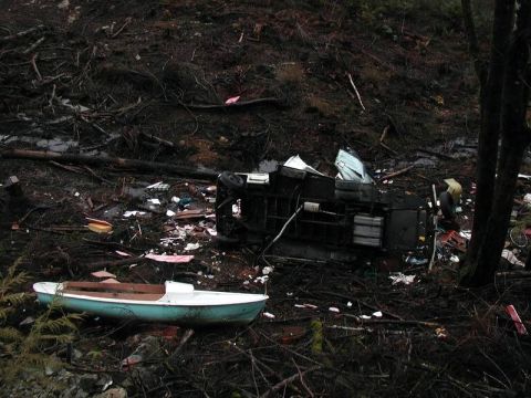 1985 Ford Bronco - A light blue and white small boat rests on the ground amidst debris and a wrecked vehicle. The boat appears to be a rowboat or dinghy with…