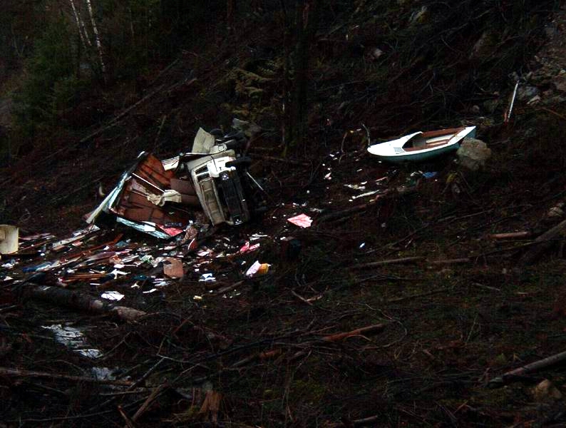 Wrecked camper and scattered debris on a wooded hillside with a small boat above.