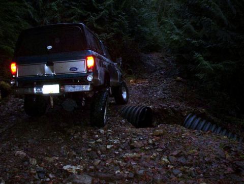 1985 Ford Bronco - The rear of a dark blue Ford Bronco is shown on a muddy, wooded trail at night. The vehicle's brake lights are illuminated, and it appears…