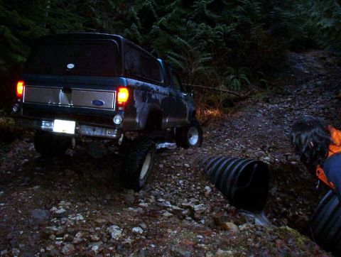 1985 Ford Bronco - The rear of a dark blue Ford Bronco is shown at night on a muddy, wooded trail. The vehicle is positioned to cross a culvert pipe with wate…