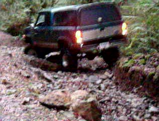 1985 Ford Bronco - A dark blue and silver SUV is navigating a rocky, off-road trail. The rear of the vehicle is visible, with its brake lights illuminated.