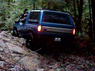 1985 Ford Bronco - A blue Ford Bronco is shown from a rear three-quarter angle, navigating a rocky, wooded trail. Its taillights are illuminated, and it appea…