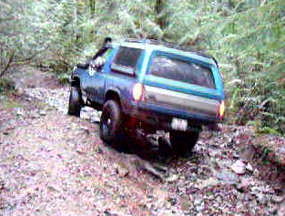 1985 Ford Bronco - A blue two-door SUV is navigating a muddy, rocky trail through a wooded area. The rear of the vehicle is angled away from the camera, showi…