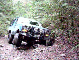 1985 Ford Bronco - A light blue 1985 Ford Bronco is shown from a front 3/4 angle, navigating a steep, rocky, off-road trail with dense foliage on the right. T…