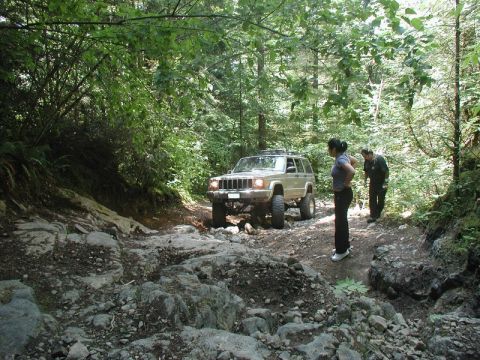1985 Ford Bronco - A tan Jeep Cherokee is navigating a rocky, wooded trail with two people standing nearby. The vehicle is positioned on a rough, uneven path…