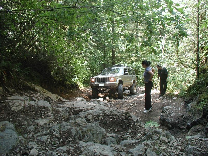 Tan Jeep Cherokee driving on a rocky forest trail with two people watching.