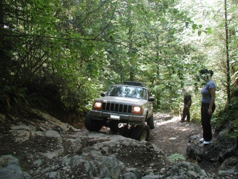 1985 Ford Bronco - A tan Jeep Cherokee is navigating a rocky, wooded trail with two people standing nearby watching.