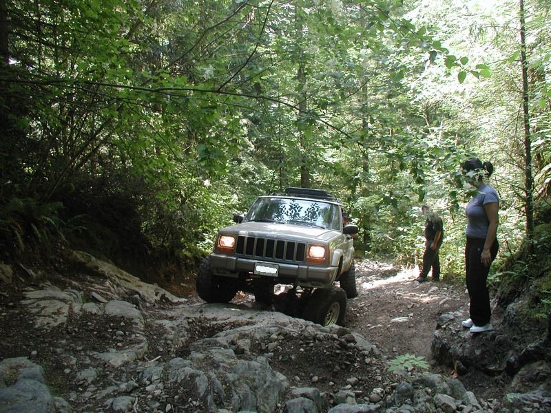Tan Jeep Cherokee off-roading on a rocky, wooded trail.