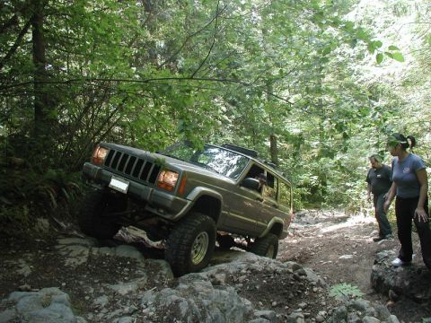 1985 Ford Bronco - A dark colored Jeep Cherokee XJ is navigating a rocky, wooded trail with two people standing nearby watching.