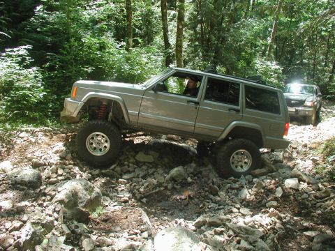 1985 Ford Bronco - A silver Jeep Cherokee XJ is navigating a rocky, wooded trail with its front passenger-side tire lifted high off the ground. Another SUV is…