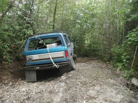 1985 Ford Bronco - A blue Ford Bronco is navigating a rocky, wooded trail. The rear of the vehicle is angled towards the camera, showing its off-road tires an…