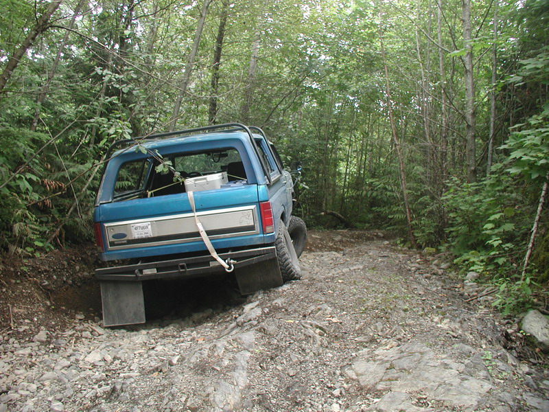 Blue 1985 Ford Bronco on a rocky, wooded trail, rear view.
