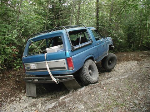 1985 Ford Bronco - A blue Ford Bronco is navigating a rocky, wooded trail with its rear end angled upwards. The vehicle has large off-road tires and a modifie…