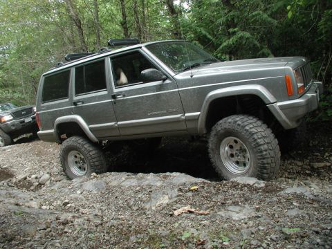 1985 Ford Bronco - A gray Jeep Cherokee XJ is navigating a rocky off-road trail, with its front passenger-side tire high in the air as it goes over an obstacl…