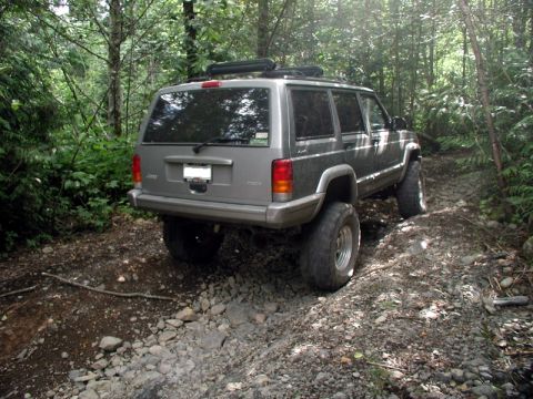 1985 Ford Bronco - A silver Jeep Cherokee is shown from the rear quarter angle, navigating a rocky, wooded trail. The vehicle has large off-road tires and a r…