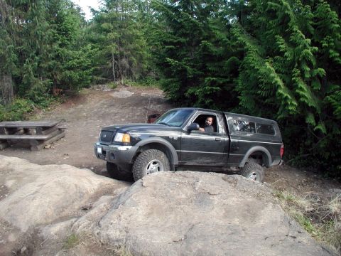 1985 Ford Bronco - A black Ford Ranger pickup truck with a camper shell is navigating a rocky, off-road trail in a wooded area. The driver is visible behind t…