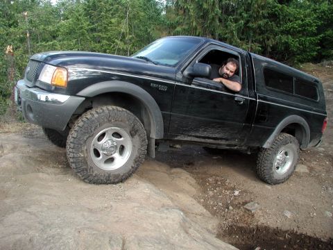 1985 Ford Bronco - A black Ford Ranger pickup truck with a camper shell is navigating a rocky, off-road trail. The driver is visible in the cab, leaning his a…