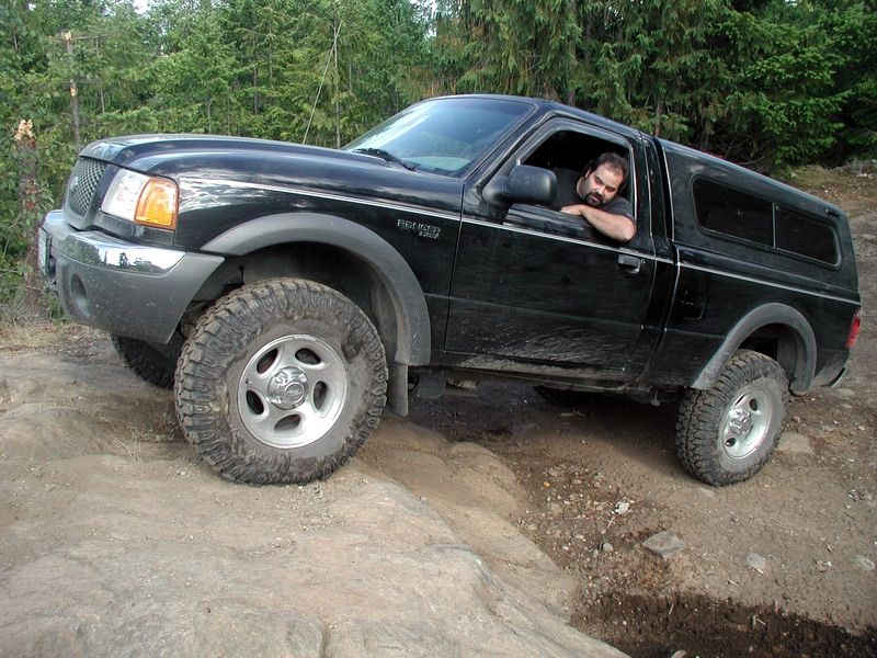 Black Ford Ranger pickup truck with camper shell off-roading on a rocky trail.