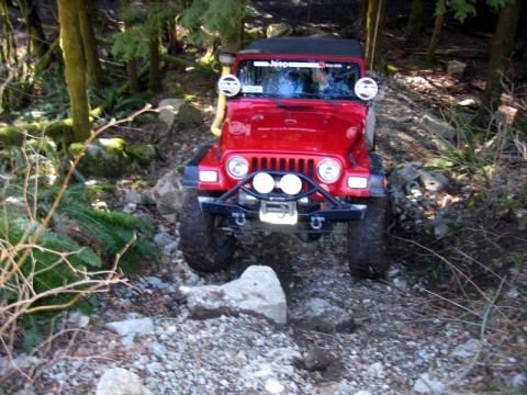 1985 Ford Bronco - A red Jeep Wrangler is navigating a rocky, wooded trail. The vehicle has a winch and auxiliary lights mounted on the front bumper.