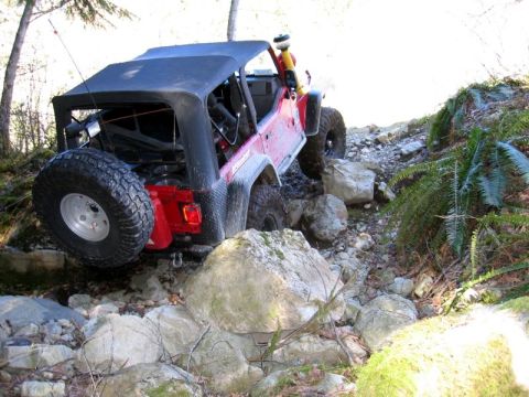 1985 Ford Bronco - A red Jeep Wrangler is navigating a rocky trail, with its rear tire elevated on a large boulder. The vehicle has a black soft top and a spa…