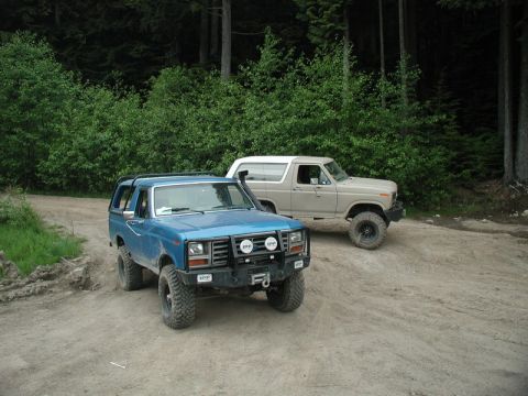 1985 Ford Bronco - Two off-road modified Ford Bronco SUVs are parked on a dirt road in a forest. The blue Bronco in the foreground has a winch and auxiliary l…