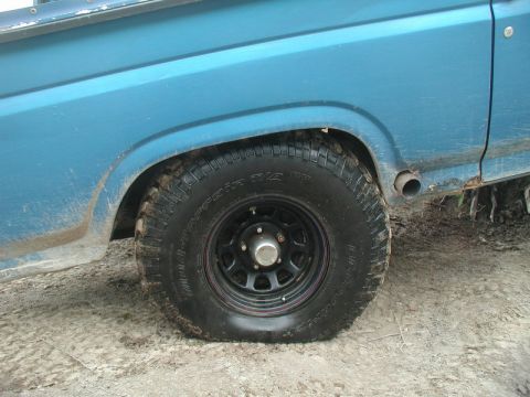 1985 Ford Bronco - A flat rear tire on a blue Ford pickup truck is shown in profile, resting on a dirt and mud surface. The tire is a BFGoodrich Mud-Terrain T…