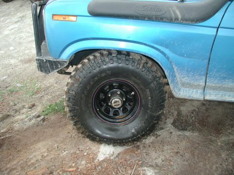 1985 Ford Bronco - The front driver's side tire and fender of a blue off-road SUV is shown, with the tire partially sunk into the dirt. The vehicle has a snor…