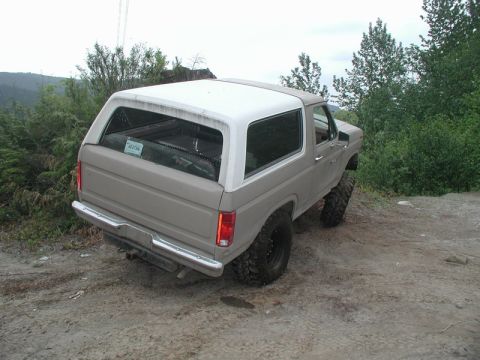 1985 Ford Bronco - The rear driver's side of a tan and white Ford Bronco is shown parked on a dirt road with trees in the background. The vehicle has large of…