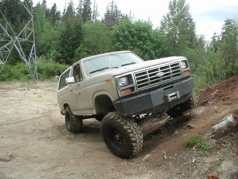 1985 Ford Bronco - A tan 1980s Ford Bronco is shown on a steep, dirt incline with large off-road tires. A large metal power line tower is visible in the backg…
