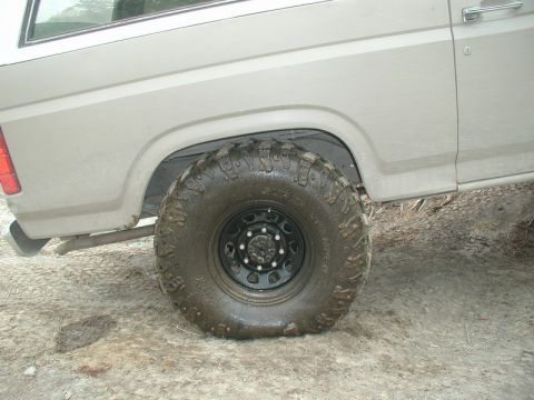 1985 Ford Bronco - The rear passenger side of a light-colored Ford Bronco is shown, with a large, muddy off-road tire and wheel. The vehicle is on a dirt and…