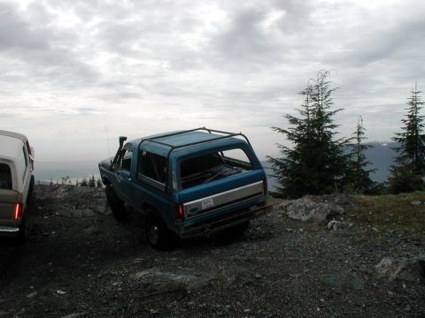 1985 Ford Bronco - A blue 1985 Ford Bronco is parked on a rocky, elevated terrain with trees and mountains in the background. The vehicle is viewed from the r…