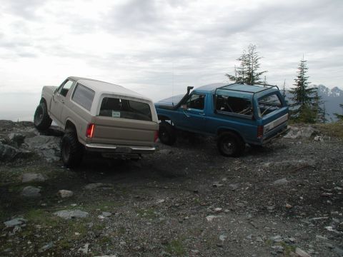 1985 Ford Bronco - Two Ford Broncos are parked on a rocky, elevated terrain with trees and mountains in the background under a cloudy sky. The Bronco on the l…